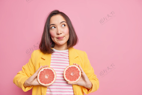 思想迷人的亚洲女人有着深思熟虑的梦幻表情抱着葡萄柚一半吃多汁的柑橘类水果燃烧热量有着健康的营养姿势靠着粉色的墙面和复制空间区思维切片沉思