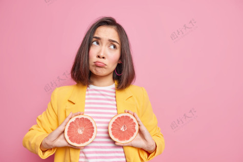 沮丧新鲜的维生素心烦意乱的亚洲女人抱着半个葡萄柚摆出悲伤的表情 美味多汁的水果摆出令人不快的姿势 穿着时髦的衣服坚持节食 隔离在粉色的墙壁空白处离开不满成人